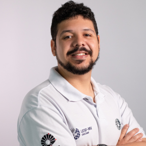 Foto colorida de homem branco de cabelos pretos curtos e barba e bigode preto. Veste camiseta estilo Polo na cor branca com o logotipo da Unicamp estampado no braço. Fim da descrição.