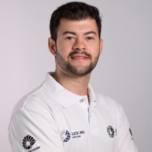 Foto colorida de homem branco de cabelos pretos curtos e barba e bigode pretos. Veste camiseta estilo polo na cor branca com o logotipo da Unicamp estampado no braço. Fim da descrição.