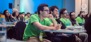 Na foto, um aluno em destaque, e vários outros em segundo plano concentrados, aparentemente prestando atenção em alguma palestra ou explicação que está sendo dada.