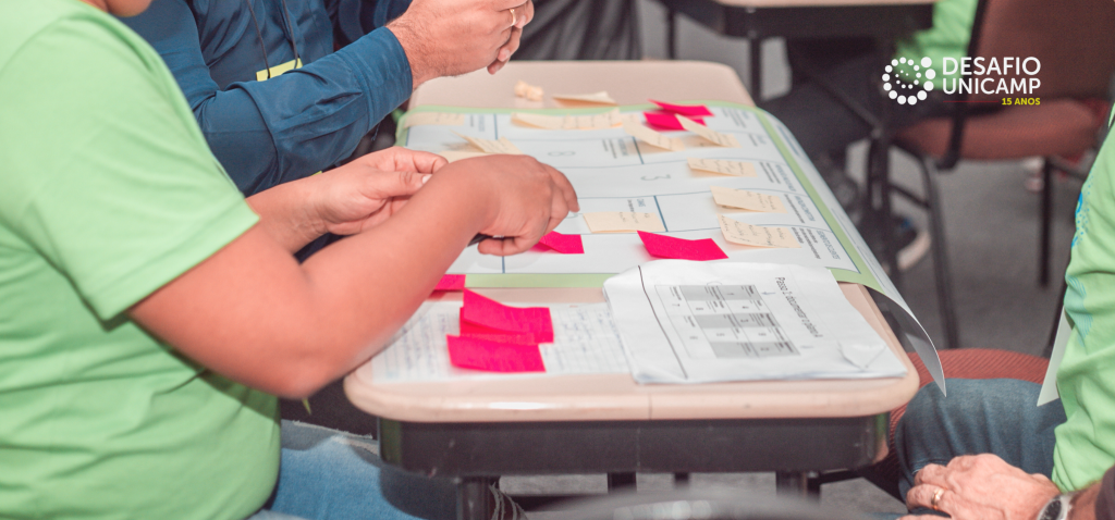 Participantes colaborando em um projeto durante a competição de empreendedorismo Desafio Unicamp, utilizando post-its e papéis para organizar ideias em grupo. Fim da descrição.