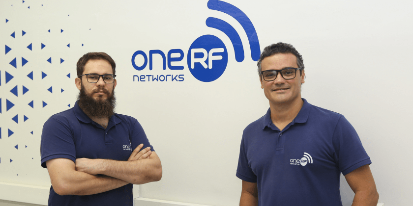 Foto posada de Gustavo Retuci e Eduardo Cardoso (esquerda para direita), fundadores da One RF. Eles encontram-se lado a lado, em frente ao logo da empresa. Eles vestem camiseta azul, com o logo da One RF. Fim da descrição.