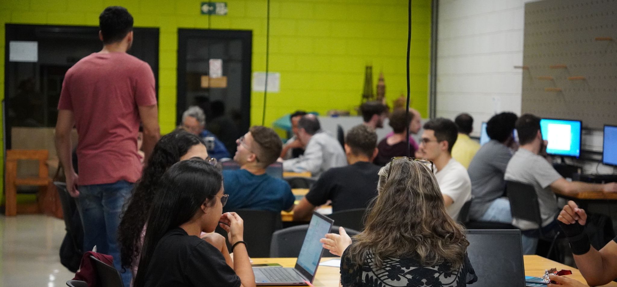 Fotografia colorida. Sala do Espaço Plasma da Unicamp, alunos da disciplina AM037 sentados nas mesas mexendo em computadores e debatendo. Fim da descrição.