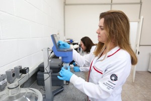 Fotografia colorida da pesquisadora, Gislaine Leonardi, uma mulher branca de cabelo loiro escuro e olhos azuis vestindo jaleco branco, mexendo em um equipamento no laboratório. Fim da descrição.