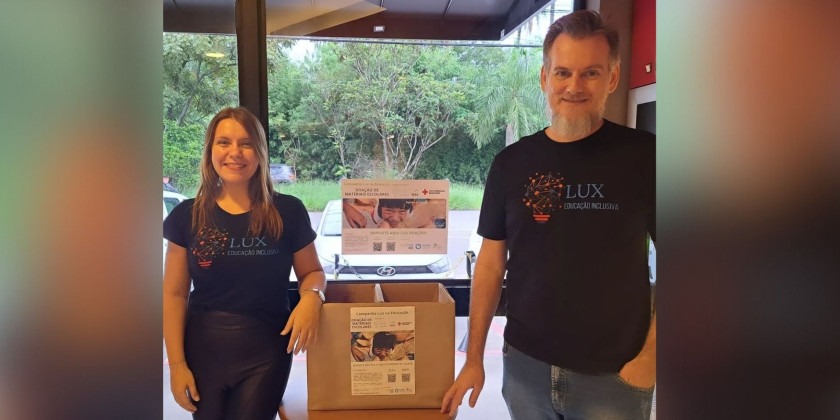 Fotografia. Os sócios da Lux Educação Inclusiva, Maibí Mascarenhas à esquerda da imagem e Paulo Kussler, à direita, com uma caixa de papelão para recebimento de doações no centro. Fim da descrição.