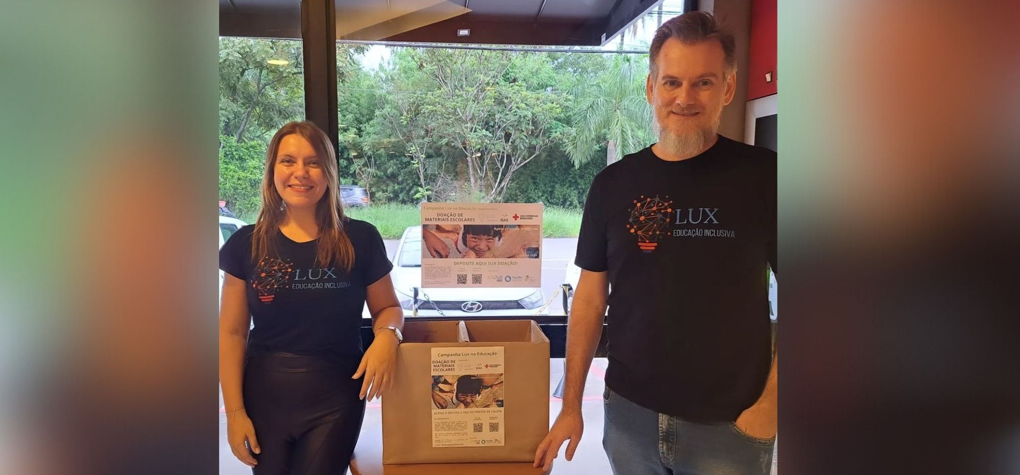 Os sócios da Lux Educação Inclusiva, Maibí Mascarenhas à esquerda da imagem e Paulo Kussler, à direita, com uma caixa de papelão para recebimento de doações no centro.
