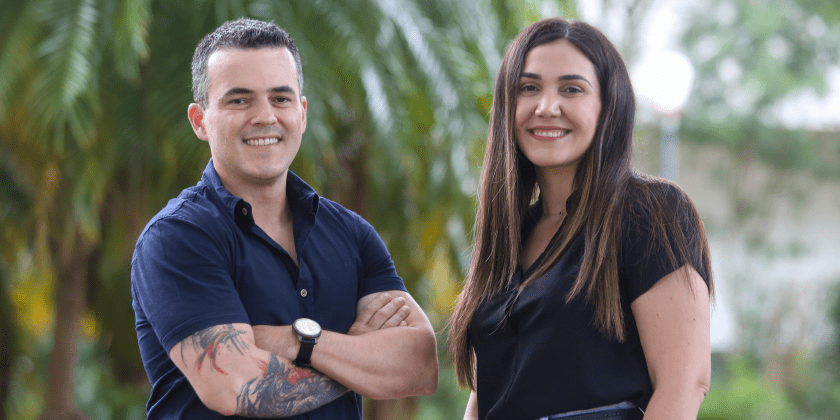 Foto posada dos fundadores da Inovia, Tiago Carvalho e Fernanda Balieiro. Na imagem, eles encontram-se olhando para a câmera, sorrindo, e contam com um árvores verdes ao fundo. O homem está de braços cruzados e veste uma camiseta azul marinho. A mulher, por sua vez, tem cabelos compridos e veste uma camiseta preta. Fim da descrição.