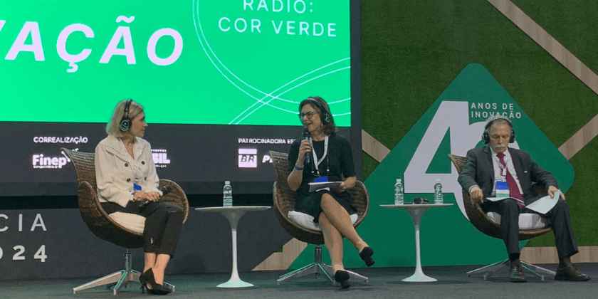 Foto colorida de palco da Conferência ANPEI de Inovação 2024 onde estão duas mulheres e um homem sentados, com fones de ouvido e microfones. A professora Ana Frattini que está ao centro, dirige uma pergunta à representante da IBM, que está a sua direita. Ao fundo há um painel luminoso verde com escritos em letras em cor branca. Fim da descrição.