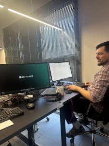 Foto colorida mostra homem branco sentado em frente a um computador. Ele está em um ambiente interno de escritório. A tela do computador mostra o logotipo da empresa. Fim da descrição.