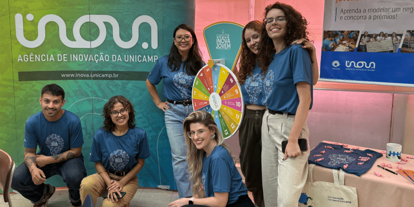 A imagem mostra seis pessoas, com a camiseta em tom azul e escrito Inova Jovem, ao redor de uma roleta e em frente a um pantográfico com o logo da Inova Unicamp. Ao lado, também há uma banner sobre o Inova Jovem, competição de empreendedorismo da Unicamp. Fim da descrição.