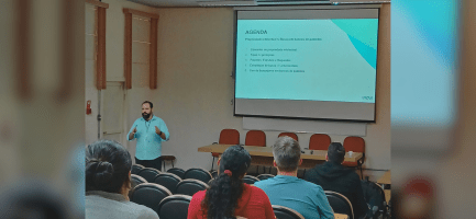 Foto tirada em ambiente interno, na Sala da Congregação do Insitituto de BIologia da Unicamp. O registro exibe o público de costas para a foto e de frente para o palestrante, que está na ponta esquerda. Há, ao fundo, um painel que exibe uma apresentação de slides. Fim da descrição.