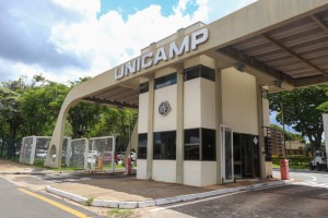 Foto externa da fachada da Unicamp, campus de Limeira. A imagem retrata a portaria da Univerisidade, que conta com o escrito "Unicamp". Ao fundo, há presença de um céu azul com nuvens brancas e algumas árvores. Fim da descrição. 