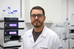 Na imagem está o professor André Damasio, do Instituto de Biologia da Unicamp e responsável pelo invento. Ele é um homem branco, com cabelo curto e castanho. Ele possui uma barba aparada e está com óculos de grau, com armação preta. A imagem mostra apenas a região do rosto e parte do tórax. Ele está vestindo um jaleco branco e olhando para a foto. Fim da descrição. 