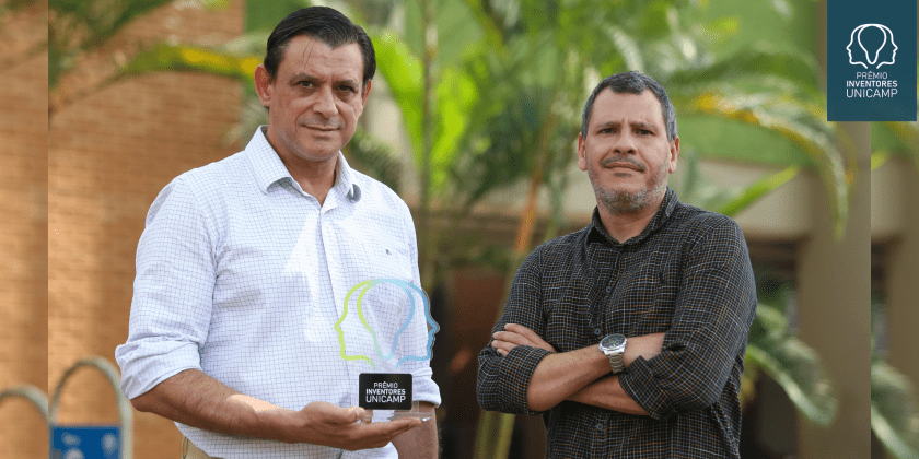 Foto colorida mostra dois homens brancos em um ambiente externo com vegetação e um edifício ao fundo. O homem à esquerda segura o troféu do Prêmio Inventores Unicamp 2024. Fim da descrição.