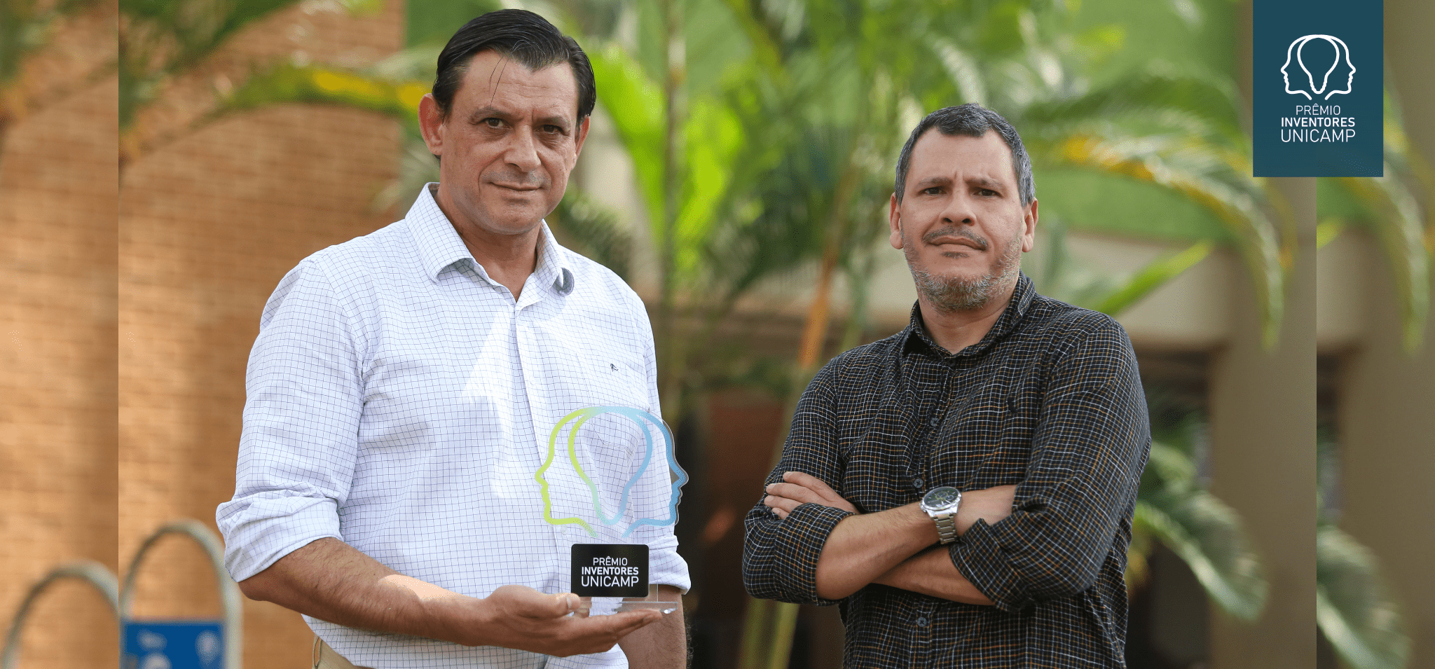 Foto colorida mostra dois homens brancos em um ambiente externo com vegetação e um edifício ao fundo. O homem à esquerda segura o troféu do Prêmio Inventores Unicamp 2024. Fim da descrição.