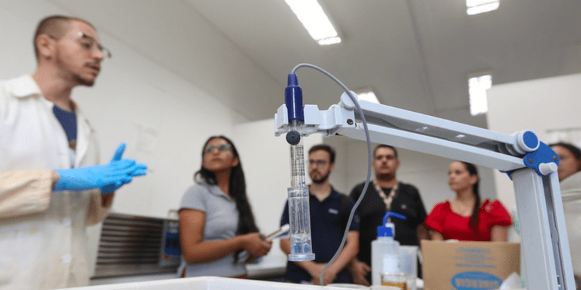 Foto de experimento em primeiro plano. Braço mecânico segurando material transparente com líquido transparente no interior. Em segundo plano desfocado há um cientista de jaleco branco e luvas azuis que explica sobre o laboratório da FEQ Unicamp para visitantes. Fim da descrição.