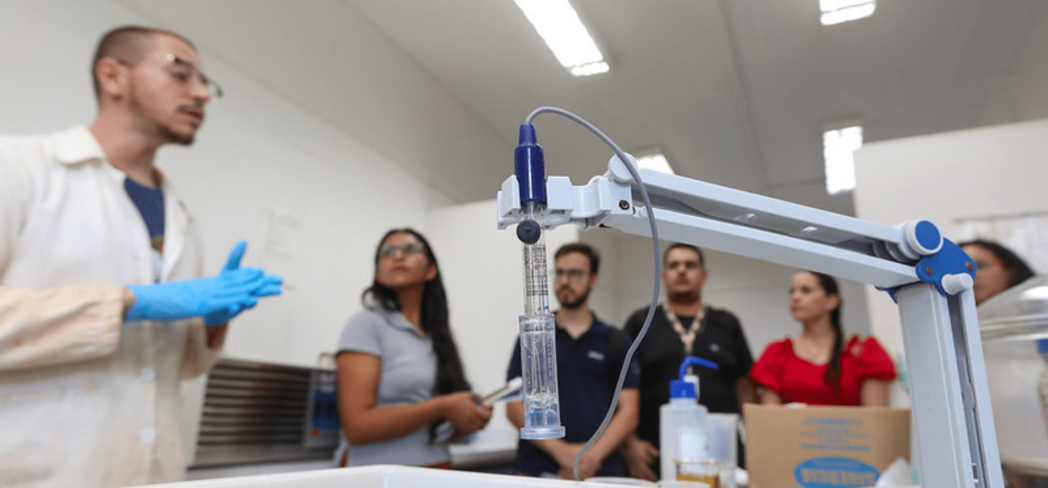 Foto de experimento em primeiro plano. Braço mecânico segurando material transparente com líquido transparente no interior. Em segundo plano desfocado há um cientista de jaleco branco e luvas azuis que explica sobre o laboratório da FEQ Unicamp para visitantes. Fim da descrição.