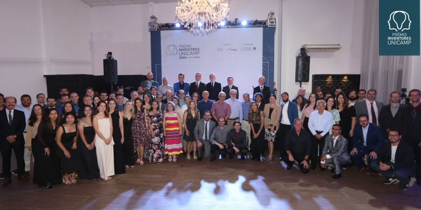 Foto dos premiados: São dezenas de pessoas lado a lado, na frente de um palco onde há um banner com o escrito Prêmio Inventores da Unicamp 2024. Os premiados estão em um salão de festas. Fim da descrição.