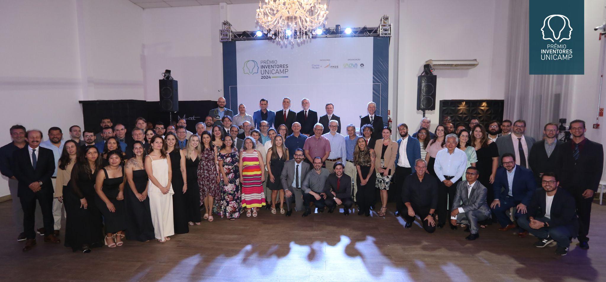 Foto dos premiados: São dezenas de pessoas lado a lado, na frente de um palco onde há um banner com o escrito Prêmio Inventores da Unicamp 2024. Os premiados estão em um salão de festas. Fim da descrição.