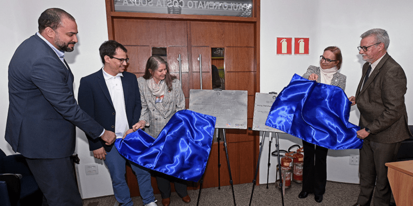 A imagem mostra o momento de inauguração do novo hub de pesquisa da Cepetro. Cinco pessoas estão presentes na foto. Eles estão retirando duas faixas que cobrem as placas de inauguração (estão "descendo a placa" de inauguração). Fim da descrição.