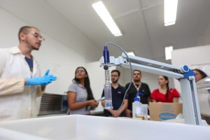 Foto de experimento em primeiro plano. Braço mecânico segurando material transparente com líquido transparente no interior. Em segundo plano desfocado há um cientista de jaleco branco e luvas azuis que explica sobre o laboratório da FEQ Unicamp para visitantes. Fim da descrição.