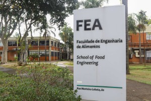  Foto colorida mostra uma placa prateada onde se lê FEA, Faculdade de Engenharia de Alimentos da Unicamp, posicionada sobre uma praça com árvores e calçamentos. Ao fundo aparece dois dos prédios da FEA.