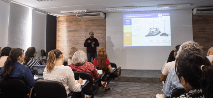 Foto feita em ambiente interno, no auditório da Incamp, traz o palestrante Clodoaldo Rulli ao fundo, ao lado de uma apresentação de slides projetada em tela. Em primeiro plano, está a plateia, de costas para o registro. Fim da descrição.