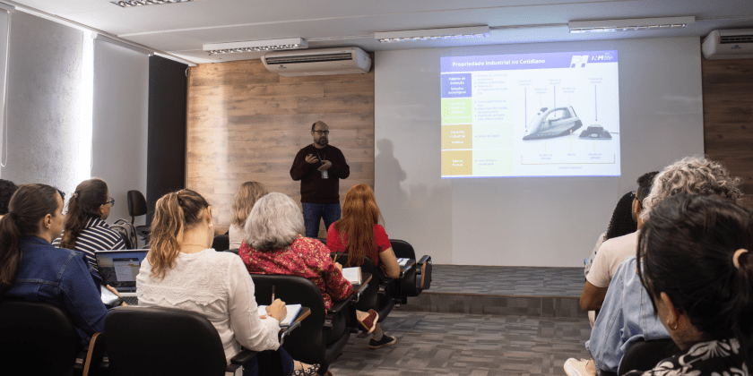 Foto feita em ambiente interno, no auditório da Incamp, traz o palestrante Clodoaldo Rulli ao fundo, ao lado de uma apresentação de slides projetada em tela. Em primeiro plano, está a plateia, de costas para o registro. Fim da descrição.