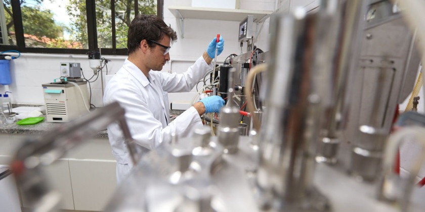 Foto interna de um homem manuseando instrumentos de laboratório. No registro, o homem encontra-se de lado para a câmera, em destaque, com elementos do laboratório em desfoque no primeiro plano. Ele veste jaleco branco, usa óculos e luvas azuis. Fim da descrição.