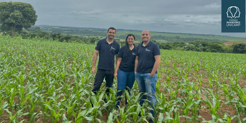 Na fotografia estão os três fundadores da startup, em pé, no meio de um campo de plantações. Eles estão vestindo uma camiseta polo na cor azul escura e com o logo da startup, escrito B.nano. Dois são homens e a que está no meio deles é uma mulher. Todos são brancos e vestem calça jeans. Fim da descrição.