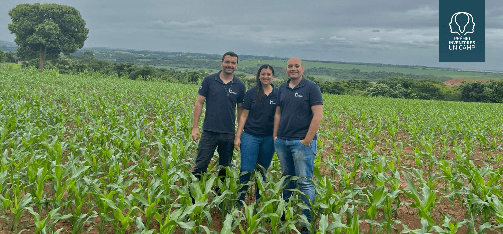 Na fotografia estão os três fundadores da startup, em pé, no meio de um campo de plantações. Eles estão vestindo uma camiseta polo na cor azul escura e com o logo da startup, escrito B.nano. Dois são homens e a que está no meio deles é uma mulher. Todos são brancos e vestem calça jeans. Fim da descrição.