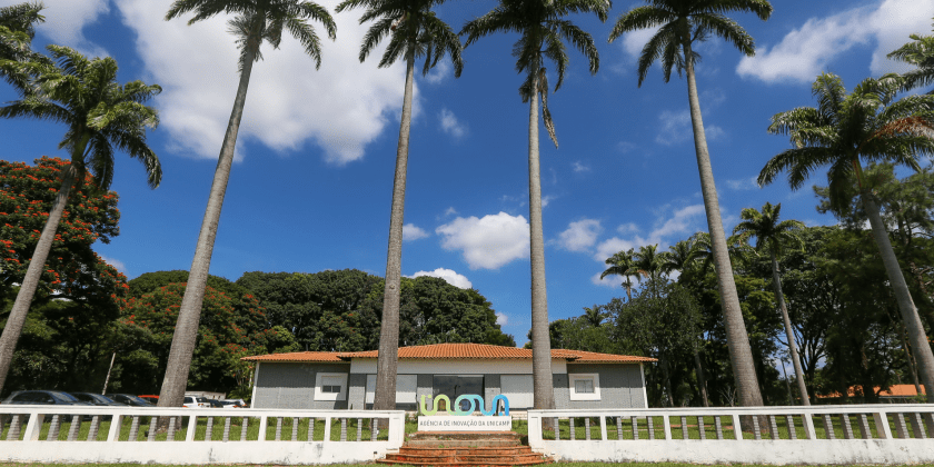 Foto em ambiente externo captura a fachada da Agência de Inovação da Unicamp. Registro conta com um totem centralizado escrito "Inova", a sede da Fazenda Argentina ao fundo, céu azul e palmeiras compondo a imagem. Fim da descrição.