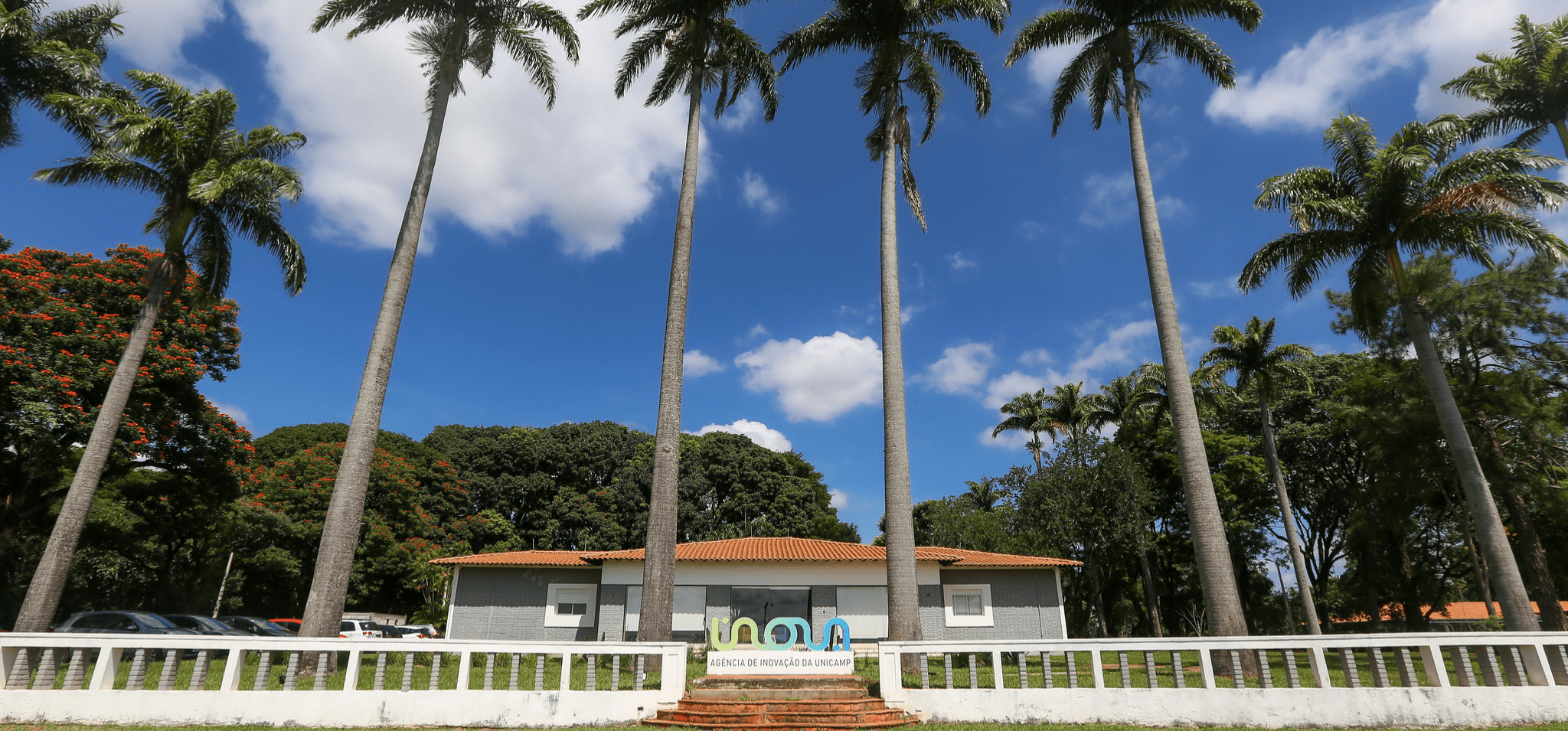 Foto em ambiente externo captura a fachada da Agência de Inovação da Unicamp. Registro conta com um totem centralizado escrito "Inova", a sede da Fazenda Argentina ao fundo, céu azul e palmeiras compondo a imagem. Fim da descrição.