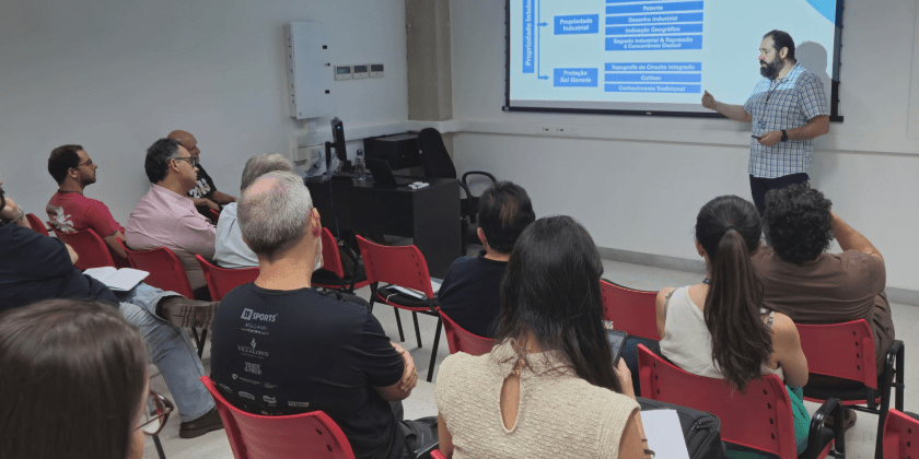 Descrição da imagem: foto em ambiente interno retratando público da oficina enquanto assistem ao palestrante. As pessoas estão sentadas de costas, de frente para o palestrante, que está ao lado de uma projeção de sua apresentação. Fim da descrição.