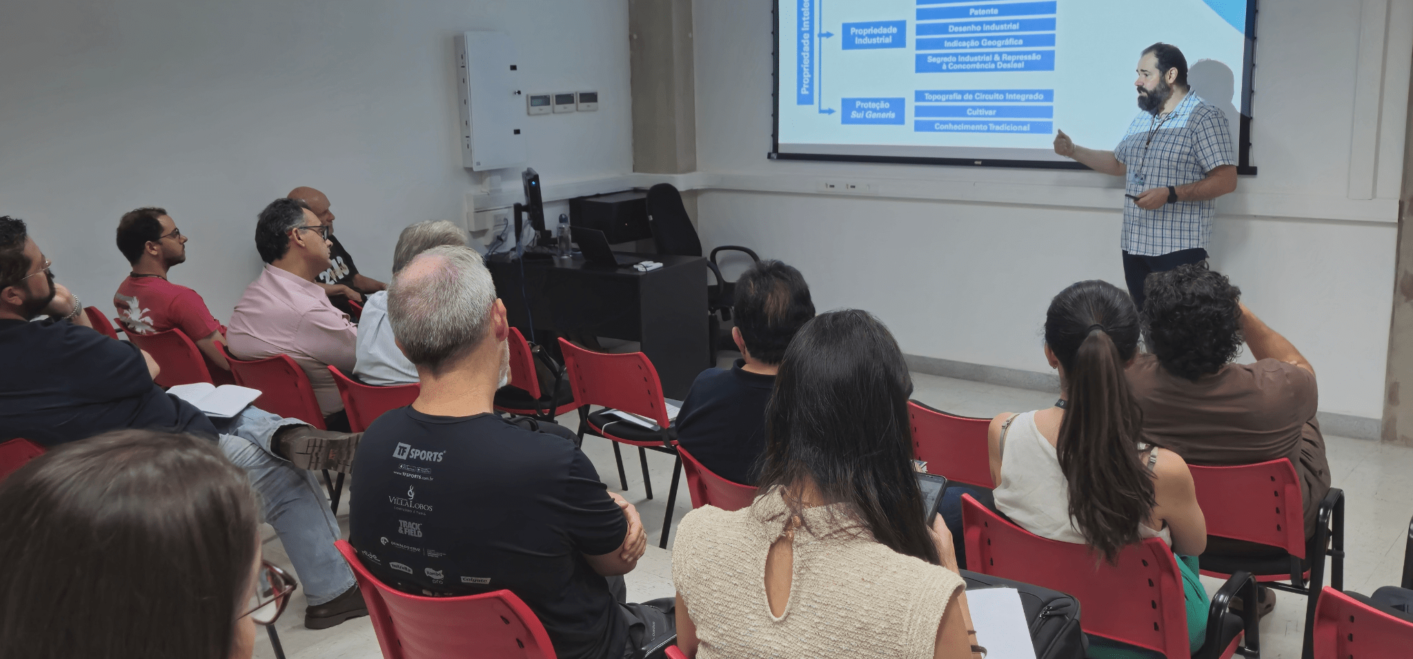 Descrição da imagem: foto em ambiente interno retratando público da oficina enquanto assistem ao palestrante. As pessoas estão sentadas de costas, de frente para o palestrante, que está ao lado de uma projeção de sua apresentação. Fim da descrição.