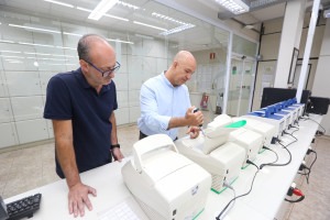 Os dois professores são homens brancos e estão juntos utilizando equipamentos laboratório. Fim da descrição.