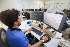 Homem branco está sentado diante de um computador em uma sala com vários outros computadores. Fim da descrição.