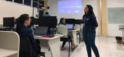 Sala de aula onde foi realizada a palestra. Duas mulheres sentadas e uma mulher em pé. Fim da descrição.