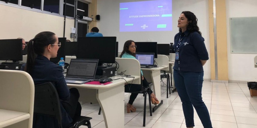 Sala de aula onde foi realizada a palestra. Duas mulheres sentadas e uma mulher em pé. Fim da descrição.