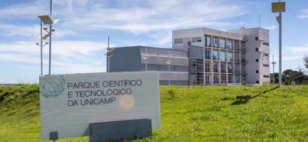 Foto externa do Parque Científico e Tecnológico da Unicamp. Imagem mostra o Prédio Núcleo, um dos edifícios que compõem a estrutura. No clique, feito em diagonal, é possível visualizar a placa indicando o nome do espaço. Há, ainda, um céu azul com nuvens brancas ao fundo e gramado verde. Fim da descrição.