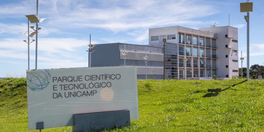 Foto externa do Parque Científico e Tecnológico da Unicamp. Imagem mostra o Prédio Núcleo, um dos edifícios que compõem a estrutura. No clique, feito em diagonal, é possível visualizar a placa indicando o nome do espaço. Há, ainda, um céu azul com nuvens brancas ao fundo e gramado verde. Fim da descrição.