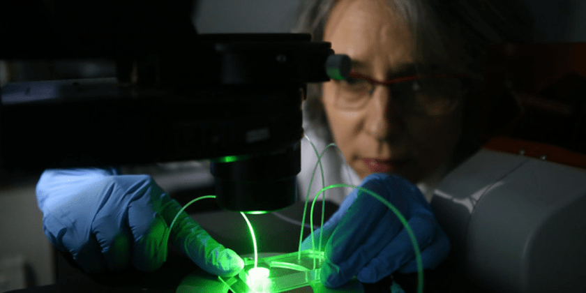 Foto de pesquisadora da Unicamp manuseando um equipamento de laboratório utilizado na produção de tecnologia que é inventora. A imagem conta com baixa iluminação e traz o equipamento em primeiro plano, com destaque para uma luz verde que sai dele. A mulher, por sua vez, está ao fundo, olhando para a ferramenta e manuseando um microdispositivo. Ela usa óculos e luvas azuis de laboratório. Fim da descrição.