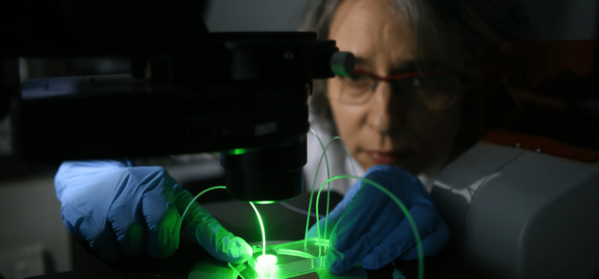 Foto de pesquisadora da Unicamp manuseando um equipamento de laboratório utilizado na produção de tecnologia que é inventora. A imagem conta com baixa iluminação e traz o equipamento em primeiro plano, com destaque para uma luz verde que sai dele. A mulher, por sua vez, está ao fundo, olhando para a ferramenta e manuseando um microdispositivo. Ela usa óculos e luvas azuis de laboratório. Fim da descrição.