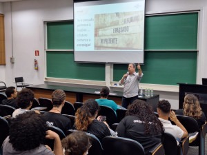 Anderson Santos, consultor do Sebrae, em segundo plano, está em pé palestrando à frente de sua apresentação. Em primeiro plano, de costas, estão alunos da Faculdade de Tecnologia da Unicamp. Fim da descrição. 