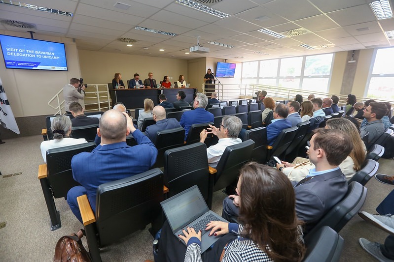 Em primeiro plano, participantes do evento. Em segundo plano, mesa de autoridades. Fim da descrição.