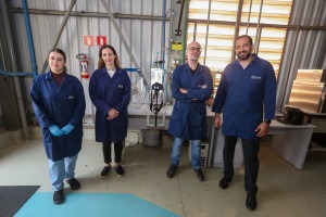 Foto de quatro pesquisadores posando em um laboratório industrial. Todos usam jalecos azuis com o logotipo "ALFA". À esquerda, uma mulher com luvas azuis está com as mãos cruzadas na frente do corpo. Ao lado dela, outra mulher também posa com as mãos cruzadas. Seguindo, um homem de óculos, barba grisalha e braços cruzados aparece ao lado de outro homem sorridente, de barba castanha, que está com os braços soltos ao lado do corpo. Ao fundo, há equipamentos de laboratório e paredes com janelas de vidro. Fim de descrição.