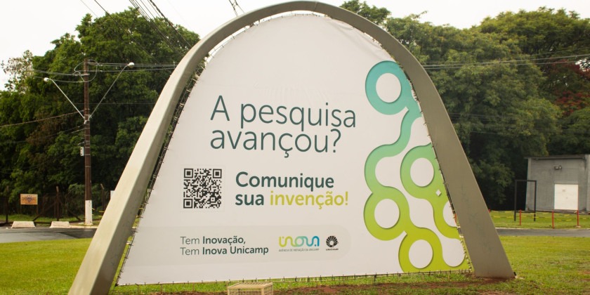 Foto de um outdoor de grande porte instalado em uma área externa, na entrada da Unicamp. O outdoor possui um design moderno, com uma estrutura arqueada e uma lona branca com impressão em alta qualidade. O texto principal diz: "A pesquisa avançou? Comunique sua invenção!". Fim da descrição.