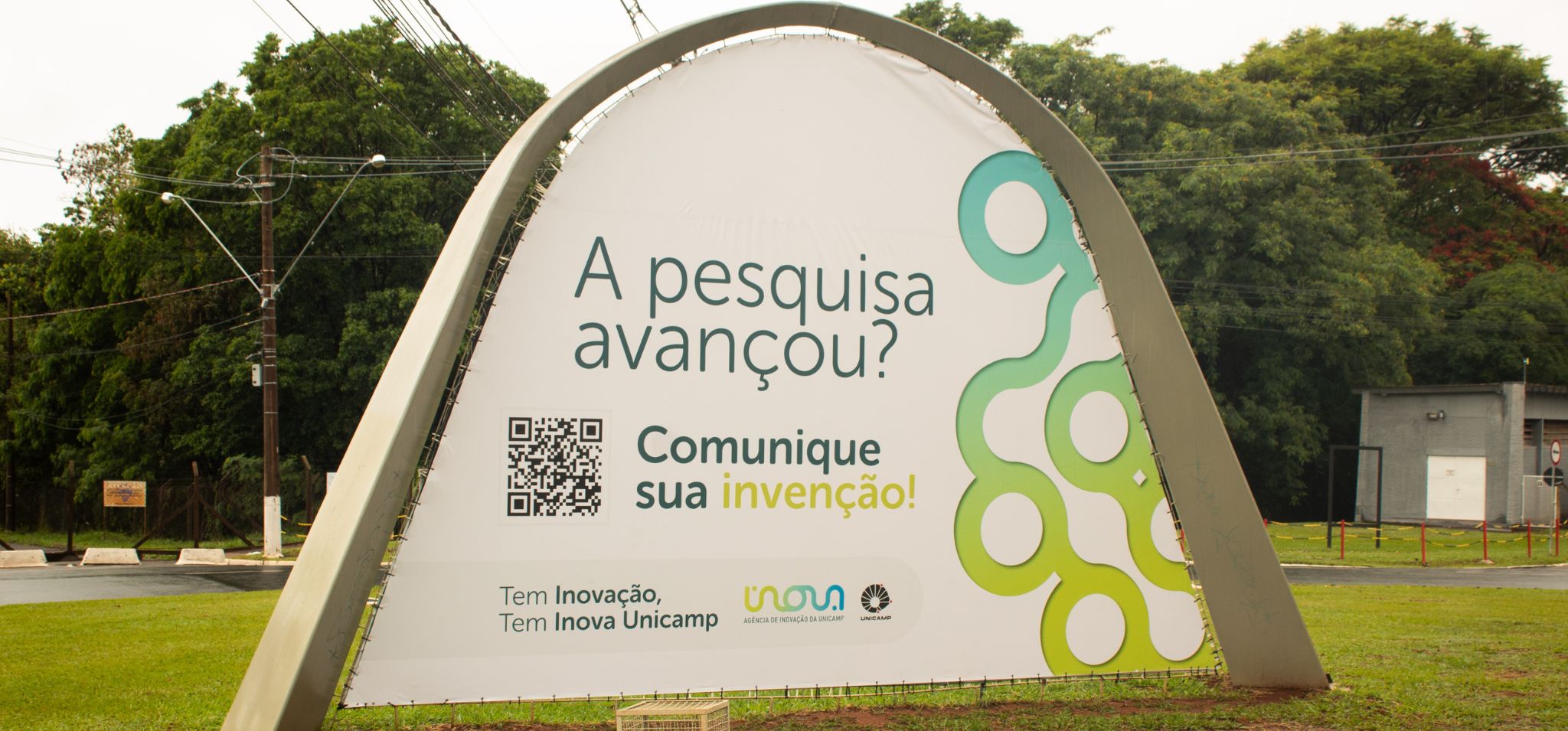 Foto de um outdoor de grande porte instalado em uma área externa, na entrada da Unicamp. O outdoor possui um design moderno, com uma estrutura arqueada e uma lona branca com impressão em alta qualidade. O texto principal diz: "A pesquisa avançou? Comunique sua invenção!". Fim da descrição.