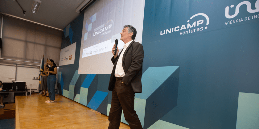 Foto colorida do professor Renato Lopes, diretor-executivo da Inova Unicamp, um homem branco de terno preto e camisa branca, que fala ao microfone. Ao fundo há um banner azul com os logotipos Unicamp Ventures, Inova e Unicamp. Fim da descrição.