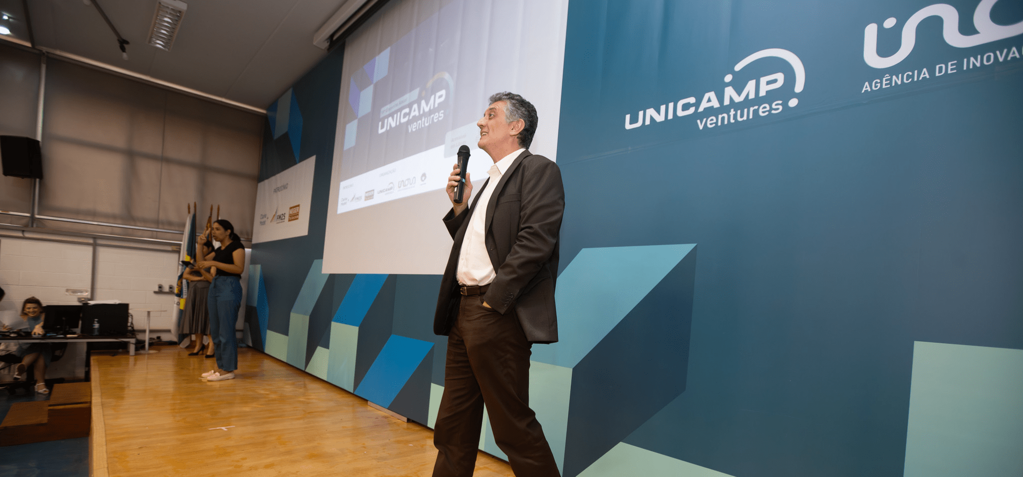 Foto colorida do professor Renato Lopes, diretor-executivo da Inova Unicamp, um homem branco de terno preto e camisa branca, que fala ao microfone. Ao fundo há um banner azul com os logotipos Unicamp Ventures, Inova e Unicamp. Fim da descrição.