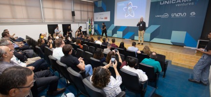 Foto colorida mostra auditório moderno com pessoas assistindo a uma apresentação no palco, onde um homem fala ao microfone. Fim da descrição.